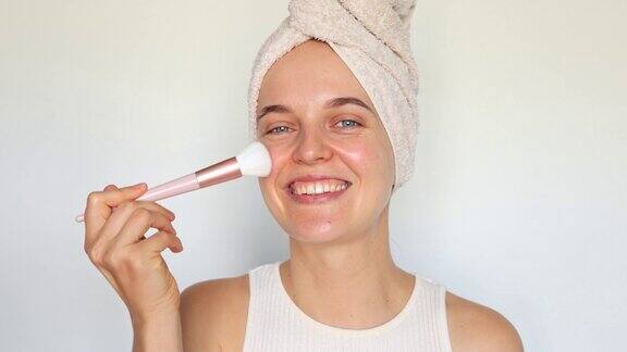 面部护理程序皮肤护理和美丽健康的年轻女子用毛巾敷在头上用化妆刷刷在脸颊上化妆淋浴后站在白色的背景上