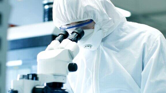 工程师技术员科学家在显微镜下检查组件的特写他在一个项目中穿着无菌工作服和防护眼镜