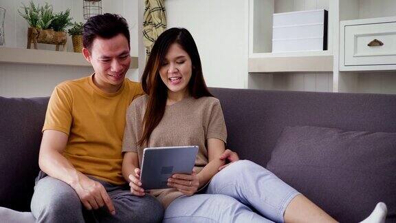 年轻的亚洲幸福夫妇坐在客厅的沙发上使用数码平板电脑夫妇们在家里放松和共度时光