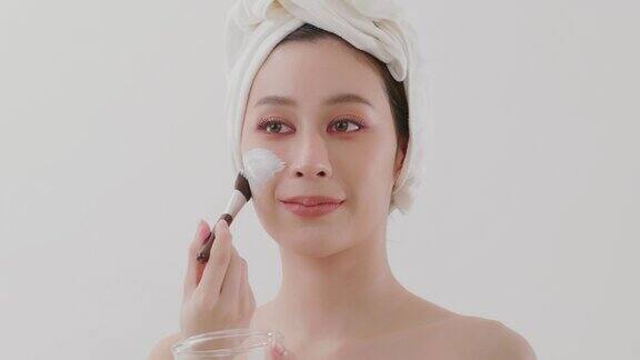 慢动作美丽的亚洲年轻女子与毛巾在她的头上应用粘土面膜在她的一半的脸使用化妆刷和镜子