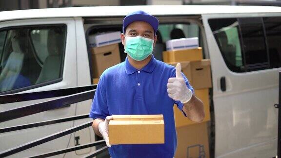 冠状病毒概念蓝色快递英俊的亚洲男子拿着包裹纸盒竖起大拇指戴着防护面具和医用橡胶手套站在货车前面慢动作镜头