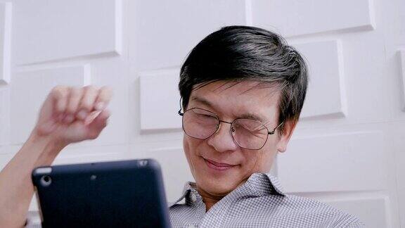 年长的亚洲男子是轻松戴眼镜拿着平板电脑使用互联网检查电子邮件坐在客厅的沙发上愉快地微笑活动生活方式概念家庭科技
