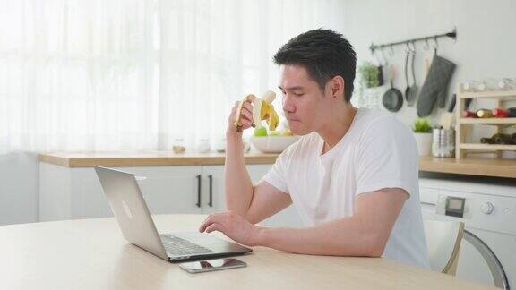 亚洲商人在家工作时吃健康的香蕉水果有吸引力的帅哥觉得开心用笔记本电脑上网学习在厨房吃干净的食物保健