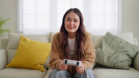 美丽年轻的亚洲女人坐在客厅沙发上拿着操纵杆遥控器玩在线视频游戏
