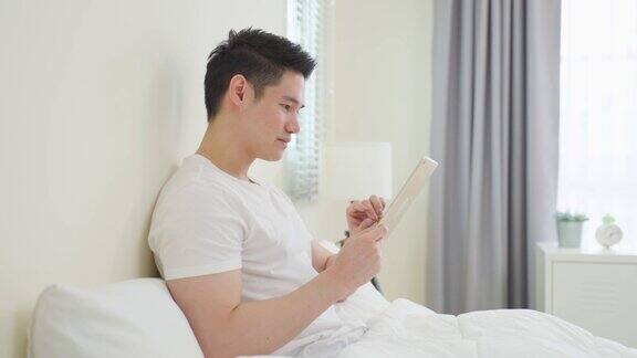 亚洲帅哥早上在家用平板电脑看网络新闻快乐有魅力的男性躺在卧室的床上享受假期周末在家里用智能手机与科技交流