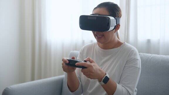 快乐的亚洲女人坐在沙发上戴着VR虚拟现实眼镜玩在线视频游戏