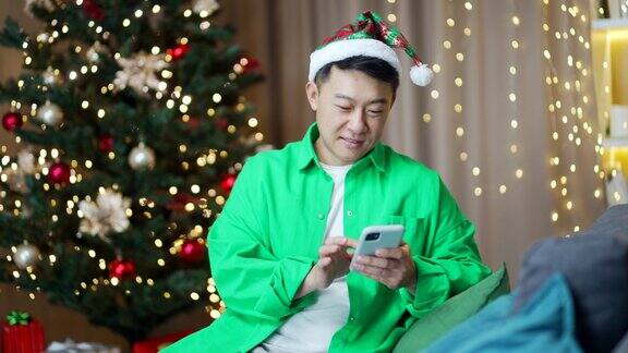 快乐的亚洲男子在新年圣诞树背景下坐在家里用手机网购输入、浏览、