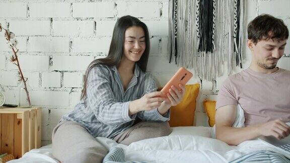 亚洲女性使用智能手机微笑显示屏幕丈夫在家床上使用笔记本电脑