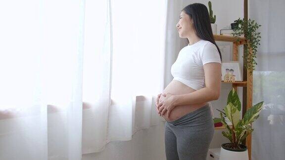 年轻的亚洲孕妇手爱抚腹部附近的窗户在家里怀孕产妇准备和预期