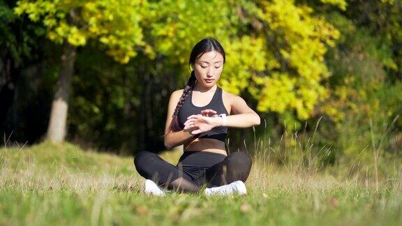 年轻漂亮的运动女孩亚洲女人在公园冥想坐莲花姿势练习瑜伽垫户外看智能手表