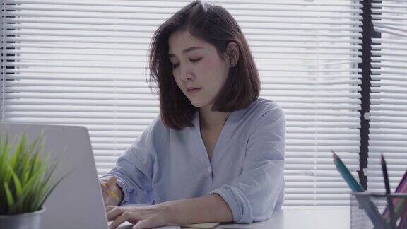 美丽的年轻微笑的亚洲女人在笔记本电脑上工作而坐在家里的客厅在家庭办公室工作的亚洲女商人享受在家的时光