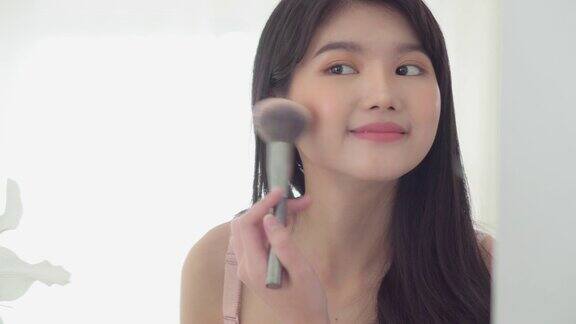 美丽年轻的亚洲女人开心地微笑着用脸照镜子在房间里用脸颊刷子化妆美丽的女孩拿着刷子在面部、皮肤护理和化妆概念上化妆