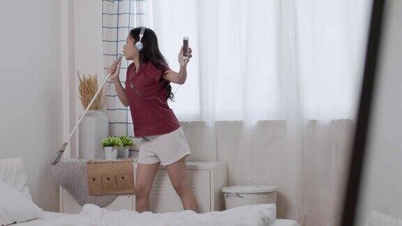 年轻的亚洲妇女戴着耳机在家里打扫卧室的时候跳舞唱歌