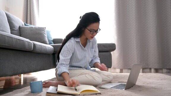 一位年轻的亚洲女商人正在笔记本电脑上寻找信息她在笔记本上做笔记她坐在家里客厅的地毯上商业女性和工作理念