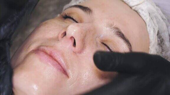 美容疗法防晒特写化妆师在黑色医疗手套应用防晒霜或均匀的化妆品霜以按摩运动女性的脸面部复兴护肤、美容、医药
