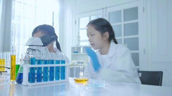 在学校的科学实验室里两个穿着大衣的可爱的亚洲女孩正目不转睛地盯着试管里的混色实验和显微镜概念教育科学实验化学