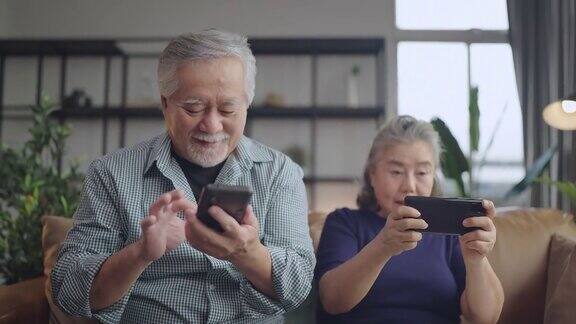 幸福亚洲退休夫妇在客厅家中室内的沙发上一起享受玩游戏和竞争游戏智能手机在线亚洲夫妇一起玩游戏家隔离的想法