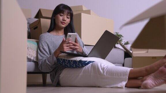 快乐的年轻亚洲女性搬到新家坐在地板上使用智能手机和笔记本电脑