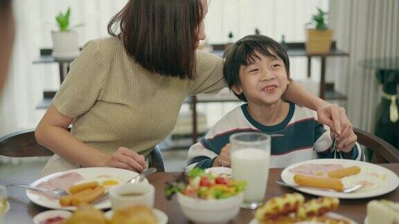 开朗的6岁亚洲小儿子和他美丽的亚洲年轻妈妈在家里吃早餐时玩得很开心小学年龄的儿子笑着吃着热狗享受在家吃饭家里做的早餐