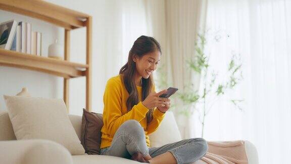 快乐的年轻亚洲女人在家里舒适的沙发上放松在智能手机上输入聊天信息微笑的女孩用手机聊天在浏览器上搜索信息无线互联网网上购物