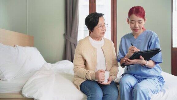 亚洲女护士穿工作服报告高级亚洲妇女的健康状况与药片在卧室护工上门拜访家庭健康护理和养老院