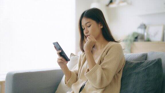 快乐的年轻亚洲女子放松在家里舒适的沙发上打字聊天信息在智能手机上微笑的女孩使用手机聊天搜索信息在浏览器无线互联网网上购物