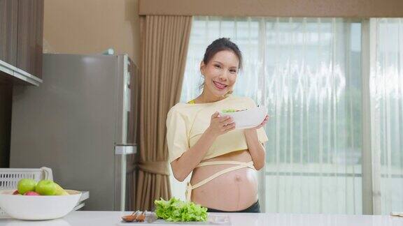 亚洲年轻孕妇的肖像拿着沙拉碗在厨房迷人的怀孕女孩站在室内感觉快乐享受吃健康的食物然后看着镜头