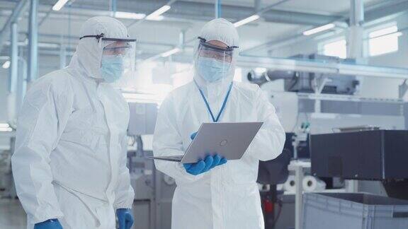 两名工程师穿着无菌工作服站在重工业工厂使用笔记本电脑检查工业制造机器设置和配置生产功能