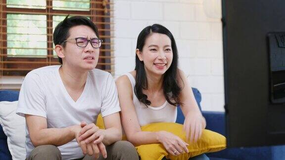 年轻的亚洲夫妇在家庭客厅的沙发上为体育节目加油助威为人们加油助威