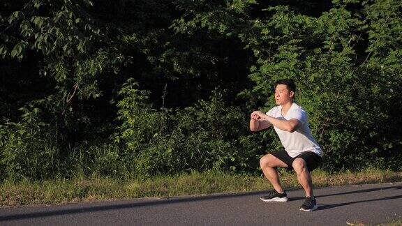 侧面视图完整的积极健康的年轻韩国男子做深蹲锻炼在街道日落的绿色树木的背景锻炼夏季男性户外运动场所运动健康
