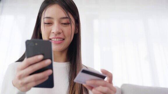 年轻的亚洲女子坐在客厅的沙发上通过智能手机上的银行卡通过互联网进行网上银行支付用信用卡在手机上网购