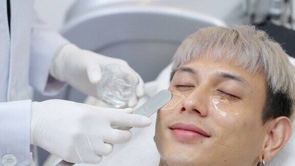 亚洲男性在美容院接受激光脱毛手术后美容师在脸上涂抹冷却凝胶