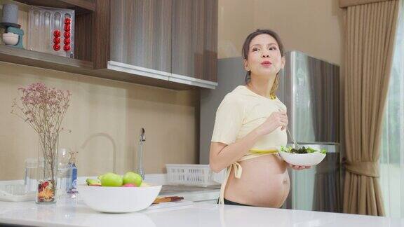亚洲美丽的孕妇在家里的厨房吃健康的食物迷人的怀孕女孩站在厨房里心情愉快享受着在碗里吃绿色蔬菜沙拉的健康