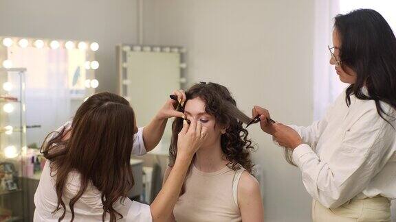 化妆师在美容工作室为漂亮的年轻女子涂睫毛膏的肖像美发师用发刷梳理年轻女子的长发