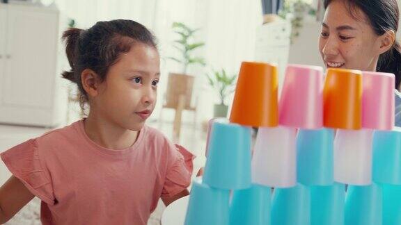 亚洲蹒跚学步的小女孩和妈妈坐在地毯上在家里客厅里玩五颜六色的叠杯玩具