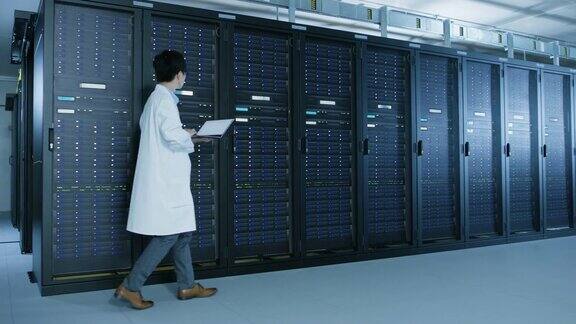 在数据中心:穿着白大褂的男性IT专家走在一排排服务器机架旁使用笔记本电脑运行维护诊断工具控制数据库平稳工作