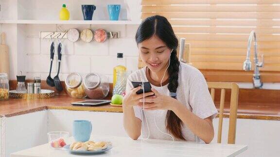 年轻美丽的亚洲女性戴着耳机用智能手机在厨房吃零食人和科技Z一代的生活方式