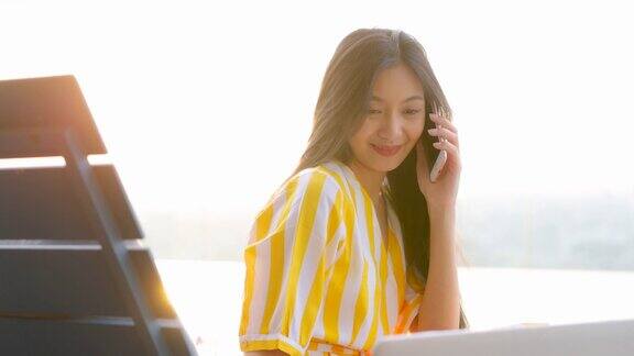 美丽的亚洲妇女在夏天黄色的连衣裙坐在游泳池附近打电话和使用笔记本电脑快乐的女顾客打电话确认网上购物订单送达