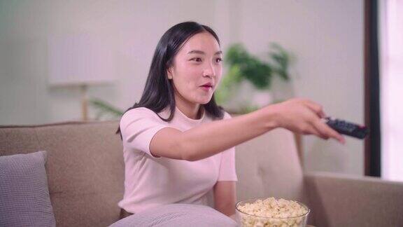 年轻的亚洲女人拿着遥控器坐在家里客厅舒适的大沙发上一边看电视一边吃爆米花