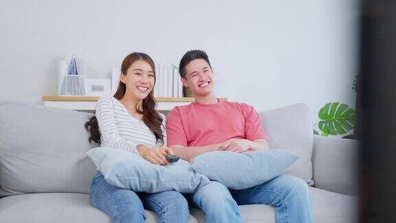 亚洲年轻情侣在家中客厅一起看电影有吸引力的浪漫的新婚姻男人和女人坐在沙发上看电视节目有乐趣的笑在一起的房子
