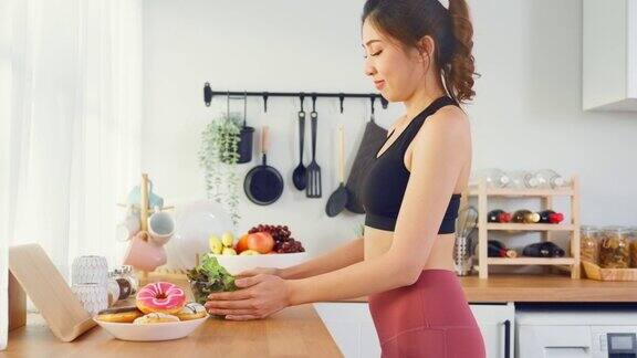 亚洲有吸引力的积极的女人拿起一个绿色沙拉在厨房在家里漂亮的运动装女孩渴望选择水果忽略甜甜甜圈享受运动后吃健康食品的健康