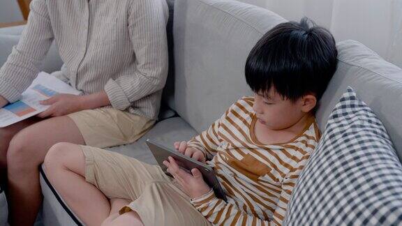 4K慢动作亚洲男孩在他最喜欢的平板电脑上玩网络游戏他高兴地坐着悬念很专注地玩游戏他坐在沙发上旁边是他美丽的单身妈妈在工作