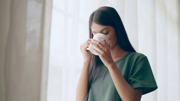 年轻的亚洲妇女站在窗边拿着杯子在卧室里她早上醒来后喝牛奶