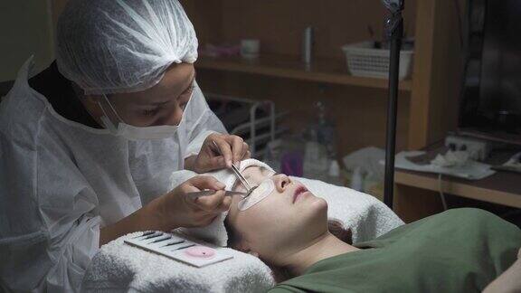 亚洲化妆师制作睫毛沙龙程序女人的睫毛扩展专业人员佩戴医用口罩帽和个人防护服在新冠肺炎疫情期间提供卫生服务年轻健康的皮肤