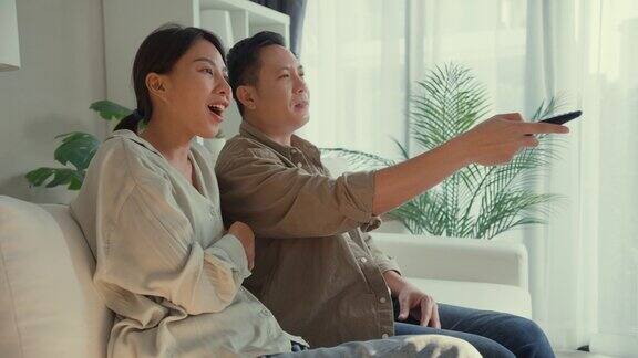 快乐的亚洲夫妇在家里的客厅坐在沙发上看喜剧频道的电影或情景喜剧生活方式花时间在家里