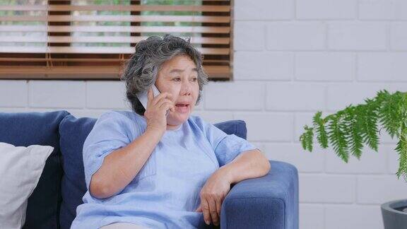 年长的亚洲女性坐在家里客厅的沙发上愉快地讲着手机老年人和科技退休生活方式