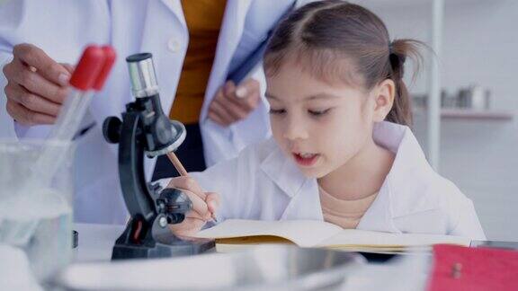 4K女学生科学室站着手拿试管前面放着小显微镜学生们正在做实验笔记听老师他们如何写下正确的实验
