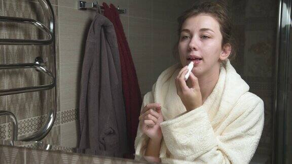 唇护理一位年轻漂亮的白人女性在嘴唇上涂上保湿卫生的口红每天化妆站在浴室的镜子旁