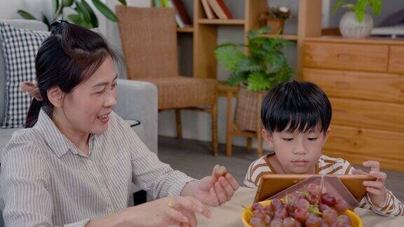 男孩坐在平板电脑上他完成了课程放松他的母亲坐在剥葡萄把种子给她的儿子并喂他的儿子一顿美味的晚餐