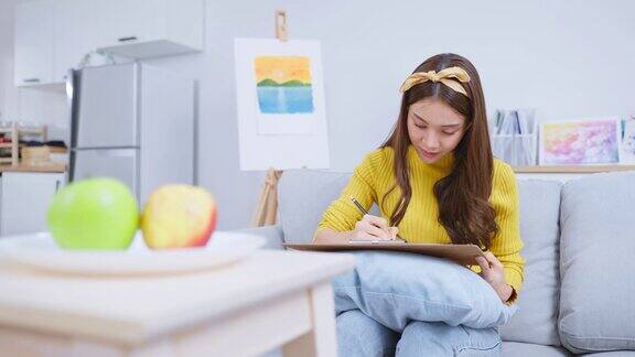 亚洲年轻有才华的女画家在画板上画画漂亮迷人的女孩坐在沙发上高兴地画出一个苹果来创作艺术品在家享受着创作活动